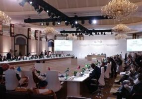 A formal conference with attendees seated around a large U-shaped table in an elegant hall.